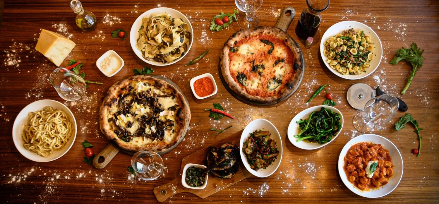 Overhead shot of pizzas, pasta and small plates on a wooden table